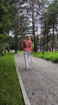 Middle aged woman walking along winding gravel path in pine forest holding white handbag and adjusting hair, peaceful summer stroll surrounded by tall trees and lush greenery captured from behind
