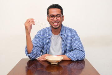 Adult Asian man holding chopsticks and sitting in front of empty dining bowl with happy face