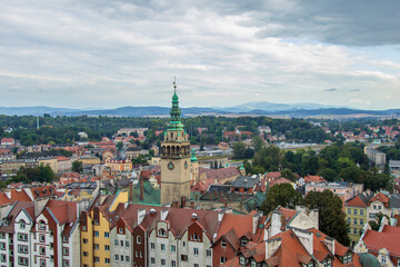Obraz premium Historic tower rising above colorful rooftops of a European town with mountain landscape in background.