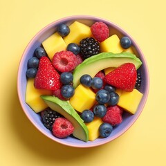 Obraz premium Colorful fruit salad with mango cubes, avocado slices, strawberries, blueberries, raspberries, and blackberries in pastel bowl on yellow background.