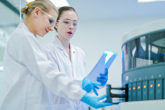 A senior scientist supervises a junior lab technician operating an automated analyzer. Two female researchers collaborating on a biomedical project in a modern high tech laboratory.