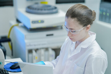 A clinical scientist documents experiment results, entering data into a computer from a printed report. An automated IHC analyzer is visible in the background of the modern pathology lab.