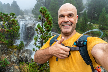 Man takes a selfie in the forest, with a watrefall on background.