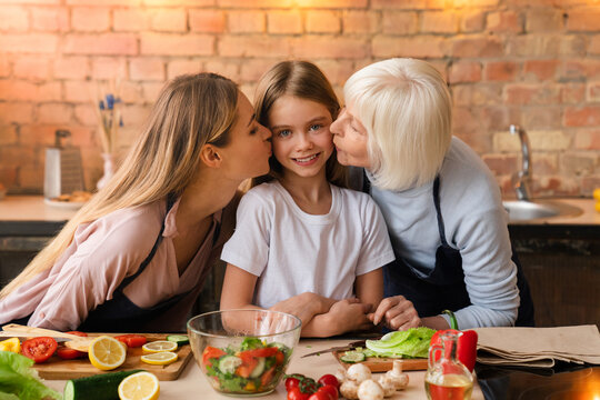 Closeup shot of little cute smiling girl that looking in camera while her mother and grandmother kissing her in home kitchen. Lovely family enjoying time with each other while cooking healthy dinner