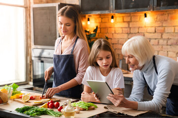 Horizontal shot of senior woman that looking in the tablet with her granddaughter and smiling. Mother cooking vegetarian dinner in kitchen. Women searching recipe in culinary website