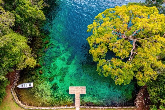 Aerial view of a tranquil cove with vibrant turquoise water, lush green trees, and a small white boat docked at a wooden pier