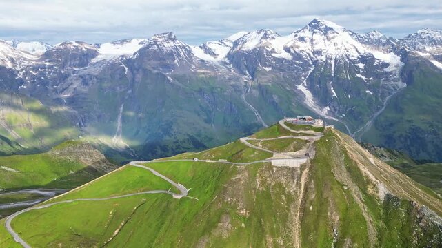 Aerial view of the winding Grossglockner High Alpine Road ascending a verdant mountain towards a structure, contrasting with snow-capped peaks, Austria.