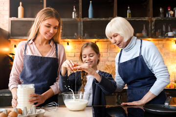 Three generations family cooking at kitchen together and having fun. Cute little girl breaks egg in in the bowl with batter. Grandmother, mother and daughter spend time together in kitchen
