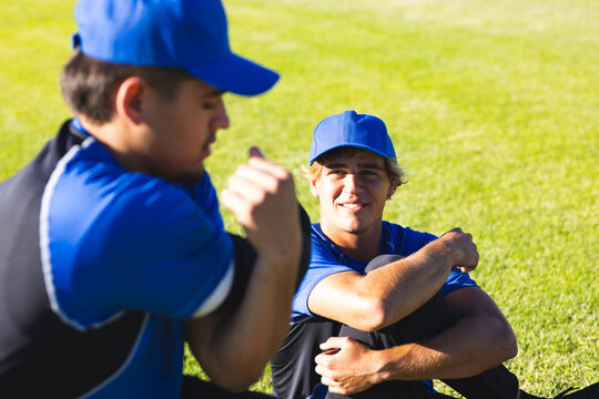 Male teammates sitting, stretching on sports field in baseball caps jerseys pants