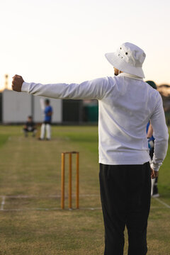 Male umpire signaling arm behind stumps and grass pitch markings with two male fielders