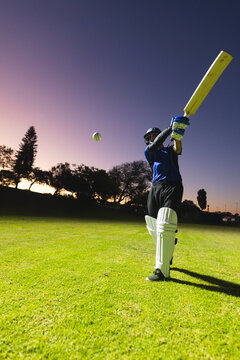 Indian male cricketer swinging bat at incoming green, white ball on grass pitch at twilight