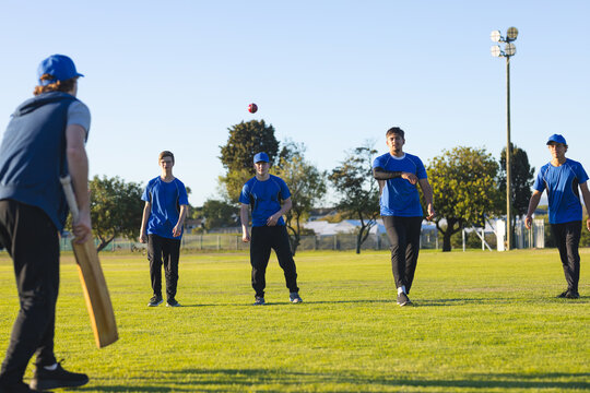 Male cricket players in blue jerseys bowling red ball and fielding on grass field