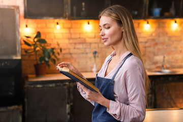 Young beautiful female caucasian woman reading book in cozy modern home kitchen while get rest of...