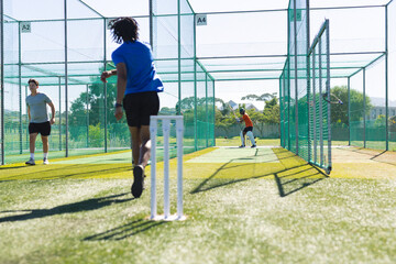 Diverse male teammates practicing bowling, batting on artificial turf in cricket net with stumps