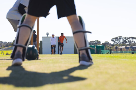 Male teammates squatting behind stumps in leg pads on field, bowler running toward pitch