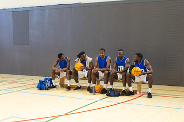 African american male teammates in blue uniforms sitting on gym bench holding basketballs