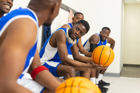 African american male partners sitting on locker room bench holding headphones, basketballs - Powered by Adobe