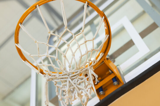 Frayed net hanging from orange basketball rim over clear backboard, in gym with beams above