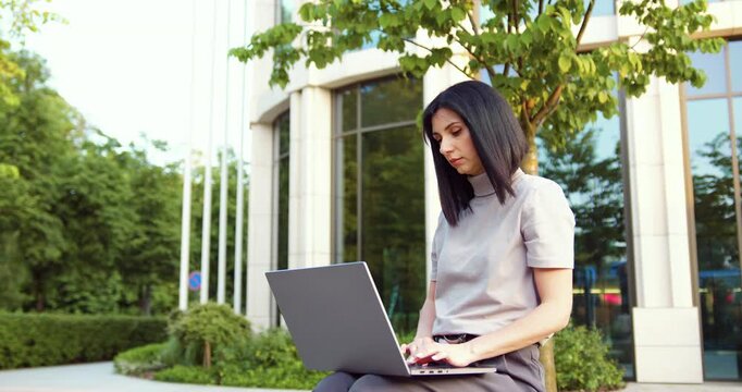 Focused young causian businesswoman using laptop pc online application for work sitting outdoors. People and Technology Concept.