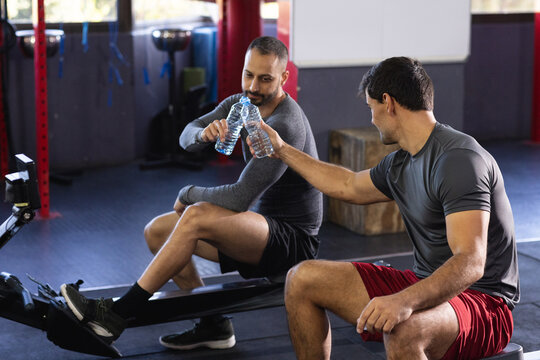 Diverse male friends sitting on rowing machines tapping water bottles next to red steel racks
