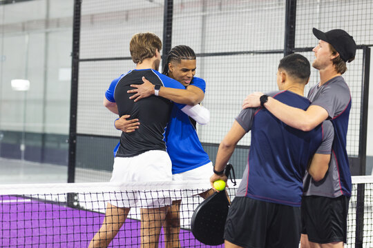 Diverse male padel teammates hugging at net on court showing wristwatches with ball and rackets - Powered by Adobe