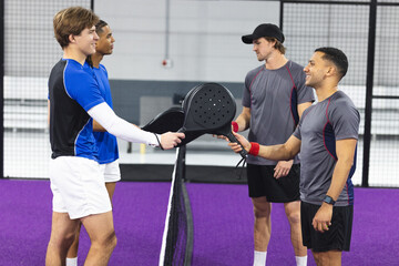 Four male amateur padel players reaching rackets across net on purple turf court with lights