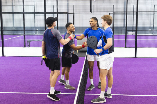 Diverse male athletes greeting on purple turf padel court near net with rackets and tennis balls - Powered by Adobe