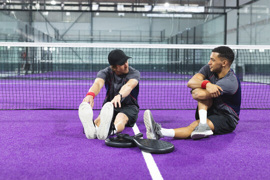 Diverse male athletes stretching on purple padel court in sportswear with rackets, wristbands