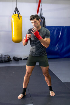 Male boxer standing in boxing stance on mat in gym with yellow punching bag