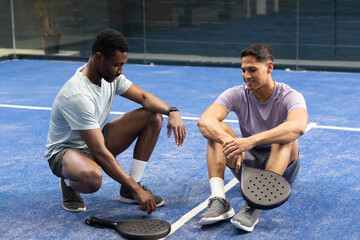 Diverse male friends kneeling, sitting on blue padel court with white lines placing down rackets
