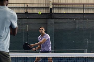 Diverse male opponents playing, tracking lime-green ball with perforated paddles on blue court
