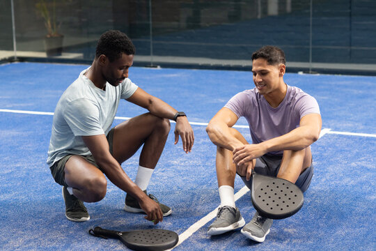 Diverse male friends kneeling, sitting on blue padel court with white lines placing down rackets