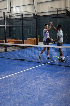 Diverse male friends practicing padel on blue court at sports arena with rackets, balls, net