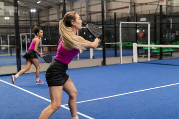 Diverse female teammates swinging padel rackets and positioning next to net on blue turf court