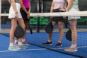 Diverse female teammates in sportswear standing at net holding black padel rackets on blue turf