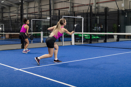 Female friends swinging padel rackets by central net on turf court with glass walls