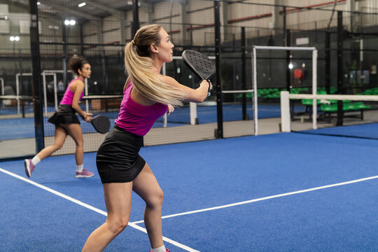 Diverse female teammates swinging padel rackets and positioning next to net on blue turf court - Powered by Adobe