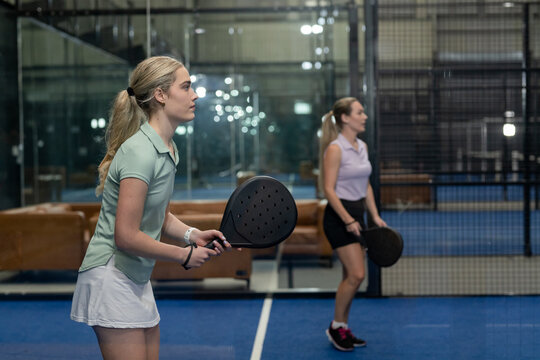 Two women playing padel with rackets on court inside glass-walled sports facility, copy space