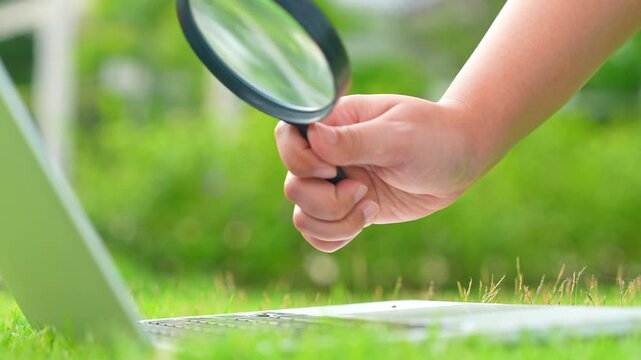 A person inspects a laptop with a magnifying glass in a green setting, highlighting digital scrutiny, curiosity, tech diagnostics, quality assurance, environmental harmony, and thoughtful exploration.