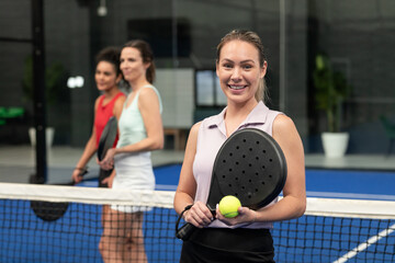Diverse female teammates standing behind padel net on court holding paddles and ball, copy space