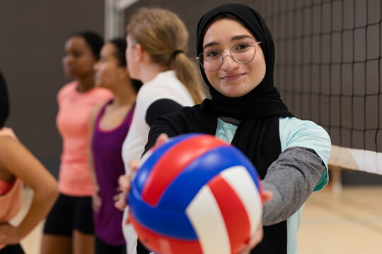 Diverse female team holding volleyball toward viewer on gym court with net, copy space