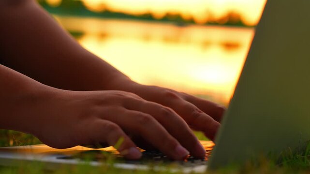 Typing on a laptop on the grass at dusk near a lake signifies digital flexibility, balanced lifestyle, serene creativity, embracing change, and blending professional and natural worlds.
