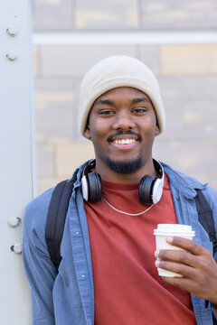African american man leaning on beam outdoors wearing backpack and headphones with coffee cup