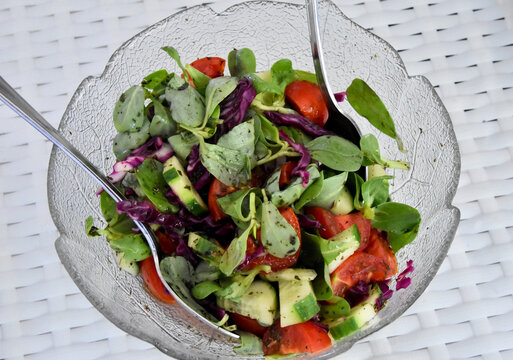 Fresh Cypriot salad in a glass bowl. A colorful, homemade salad with tomatoes, cucumbers, red cabbage, and fresh purslane leaves in a clear glass bowl, with two serving spoons.