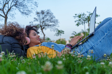 Woman with curly hair lying on grass using laptop and headphones in park