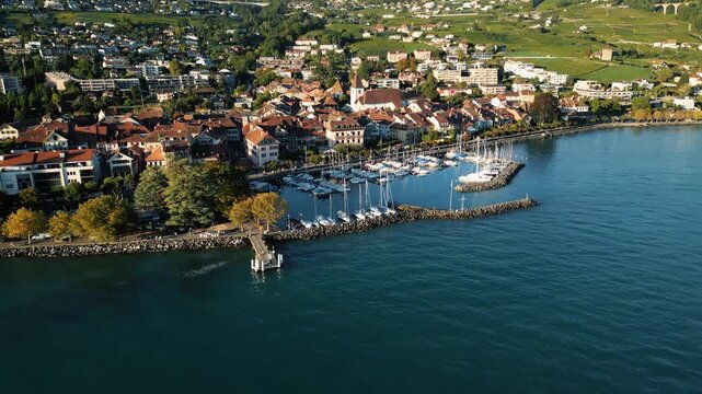 Aerial view of Lutry's marina, with boats docked along the shore, and the town's architecture blending with the vibrant blue of Lake Geneva, Lutry, Vaud, Switzerland.