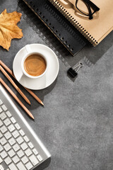 An autumn desktop with a keyboard, coffee cup, notebooks, pencil, glasses, binder clip and scattered fall leaves on a gray desk surface.