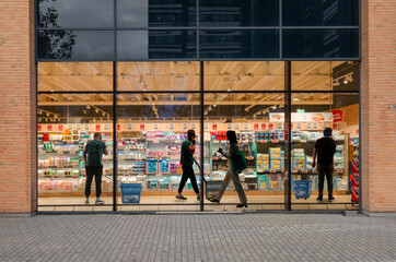A look through a window in a supermarket at day time with people with people walking around shopping, looking at goods and talking on their mobile phones.