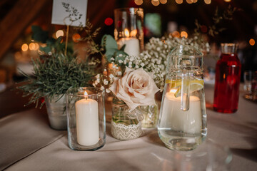 Close-up of a romantic wedding table centerpiece with candles, flowers, herbs, a water jug with lemon slices, and a red drink on a neutral tablecloth.