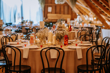 Elegant rustic wedding reception setup with round tables, floral centerpieces, candles, glassware, and wooden chairs in a warmly lit venue.